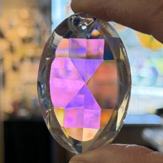 Faceted oval crystal gem held between two fingers, showing iridescent pink and purple rainbow reflections in light.