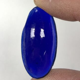 Oval cobalt blue glass cabochon gemstone held between fingertips (thumb and index finger).